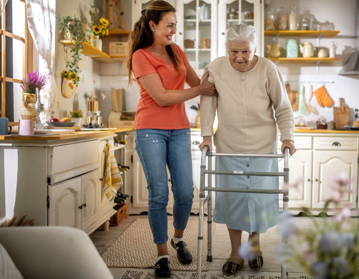 A support worker walking alongside an older woman using a walking frame in her kitchen at home