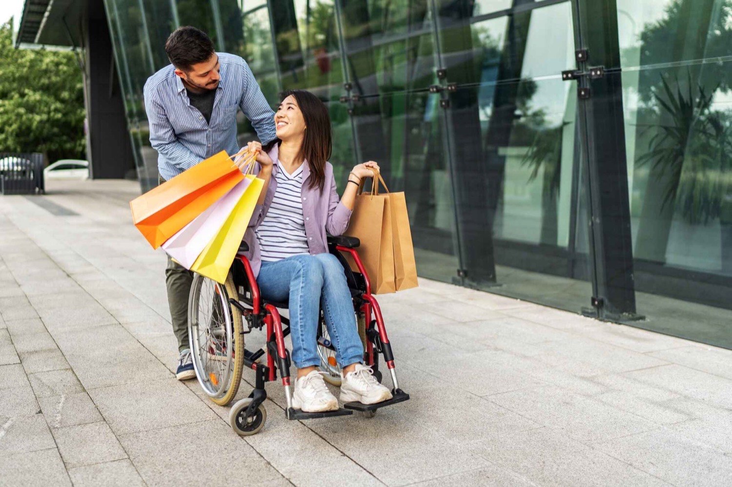 A support worker walking alongside a woman using a wheelchair, both carrying shopping bags on a city street
