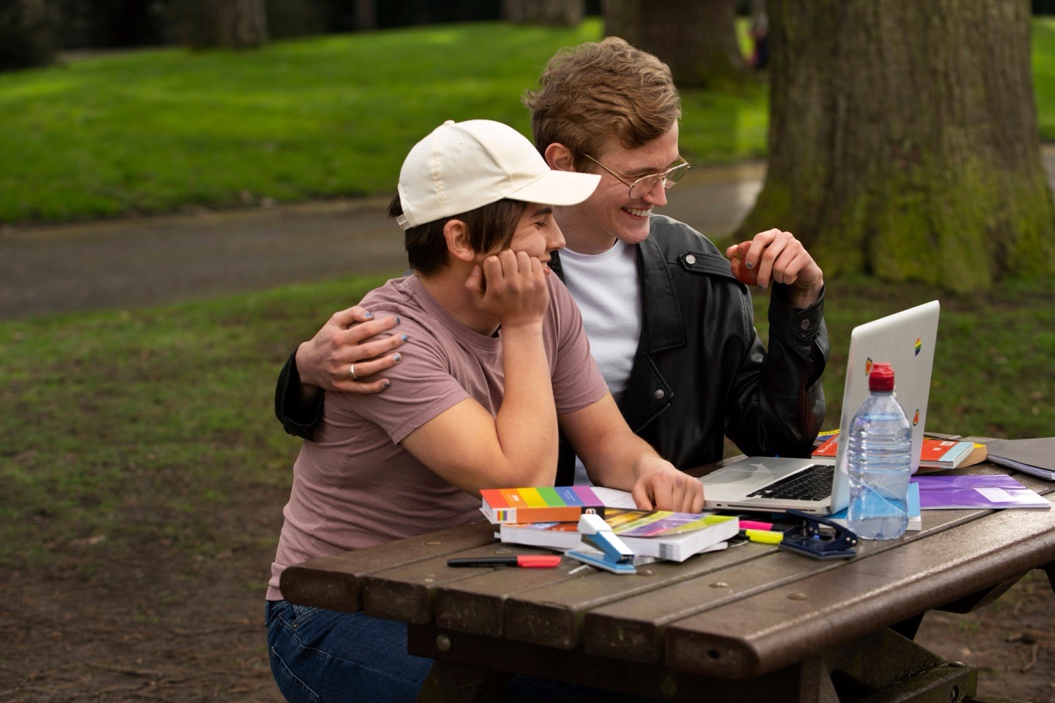 Two people sitting together at an outdoor table, one supporting the other while they work on a laptop